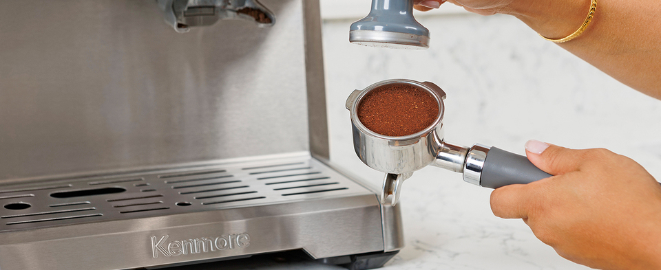 The picture shows a person holding the portafilter filled with ground coffee in one hand and the tamper in the other.