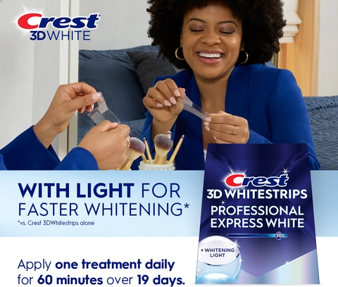 Reflection of woman in mirror smiling and peeling a Crest 3DWhitestrip. With Light for Faster Whitening.