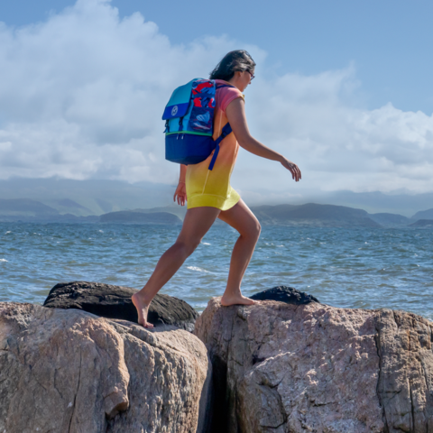Woman wearing cooler as backpack walking on the rocks by the water.