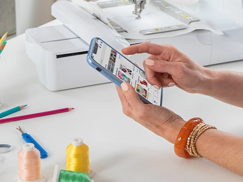 woman's hand, holding a cellphone -using Artspira App. Sewing machine on a table, with threads, pencils and sewing tool