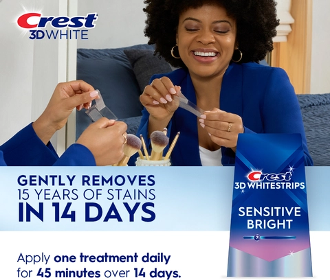 Reflection of woman in mirror smiling and peeling a Crest 3DWhitestrip. Gently Removes 15 Years of Stains in 14 Days..