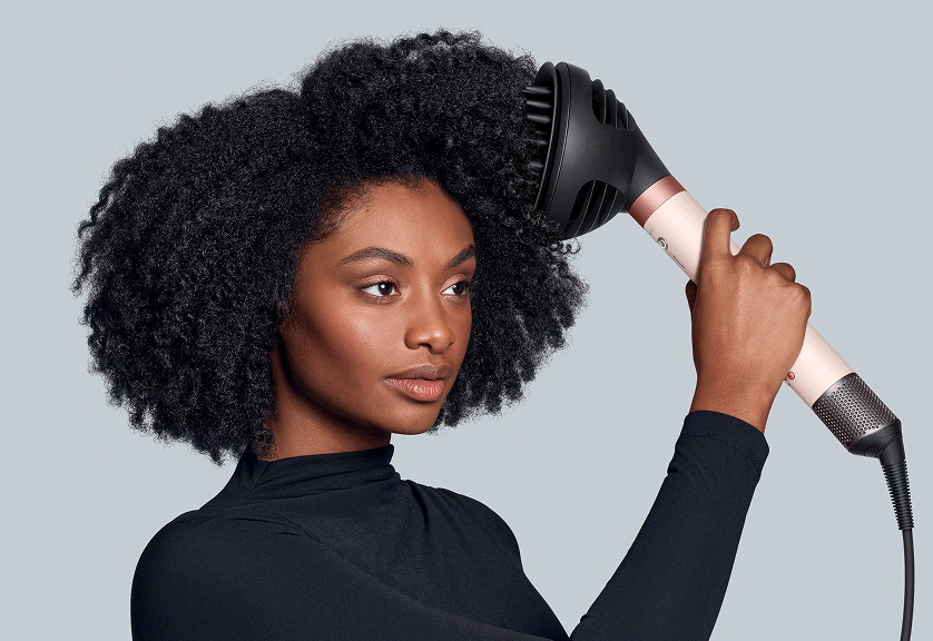 The Dyson Airwrap Co-anda2x multi-styler and dryer with the Wave+Curl diffuser 2x used by a model with dark coily hair.