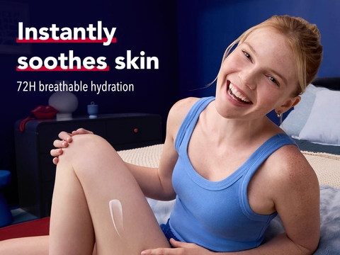 Woman applying Aquaphor lotion to her leg, smiling in comfortable bedroom with text 'instantly soothes skin