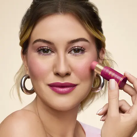 Imagem da Niina, uma mulher branca de cabelo castanho que está preso, segurando o produto Blush Stick Daily próximo ao rosto, como se estivesse aplicando-o na região das bochechas.