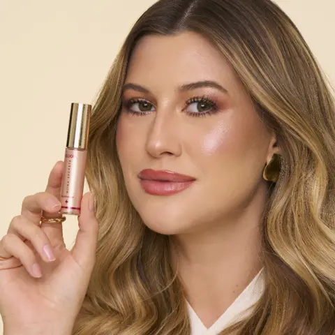 Imagem da Niina, uma mulher branca, com cabelo castanho levemente ondulado, segurando o Iluminador Luminous próximo ao rosto.