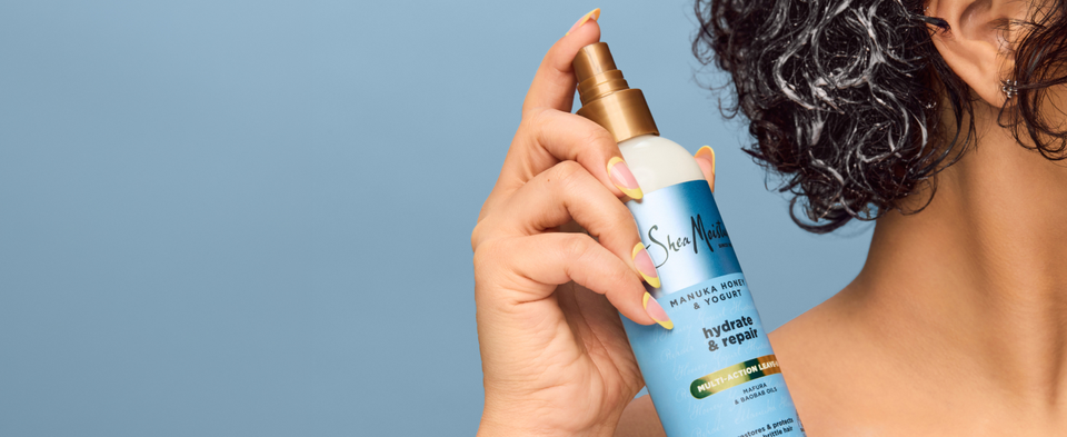 Hand spraying SheaMoisture Multi-Action Leave-In onto damp curls on a light blue background.