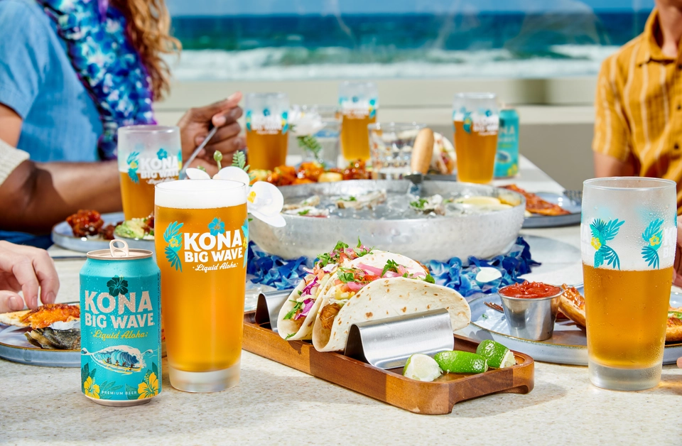 Friends gathered around a table of food on the beach, taco platters and glass of Kona Big Wave Premium Beer.