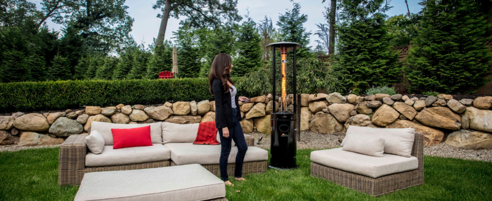 Woman standing next to Illume Propane Patio Heater in lush backyard with outdoor furniture. Woman is holding a remote control.