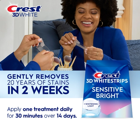 Reflection of woman in mirror smiling and peeling a Crest 3DWhitestrip. Gently Removes 20 Years of Stains in 2 Weeks.