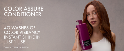 Young woman with vibrant wavy red hair holding a Nexxus Color Assure Conditioner bottle in one hand.