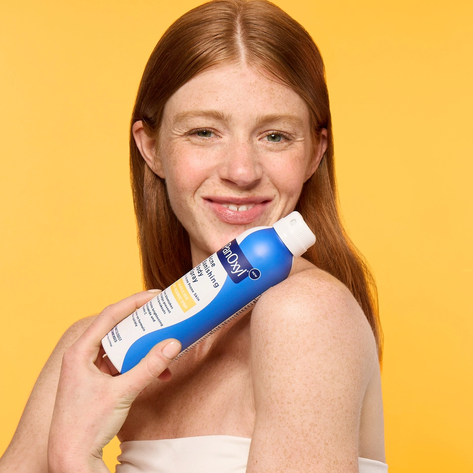 Young female holding the PanOxyl Acne Banishing Body Spray against a yellow background.