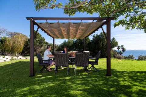 Couple sitting under the Modena pergola in a lush green backyard.