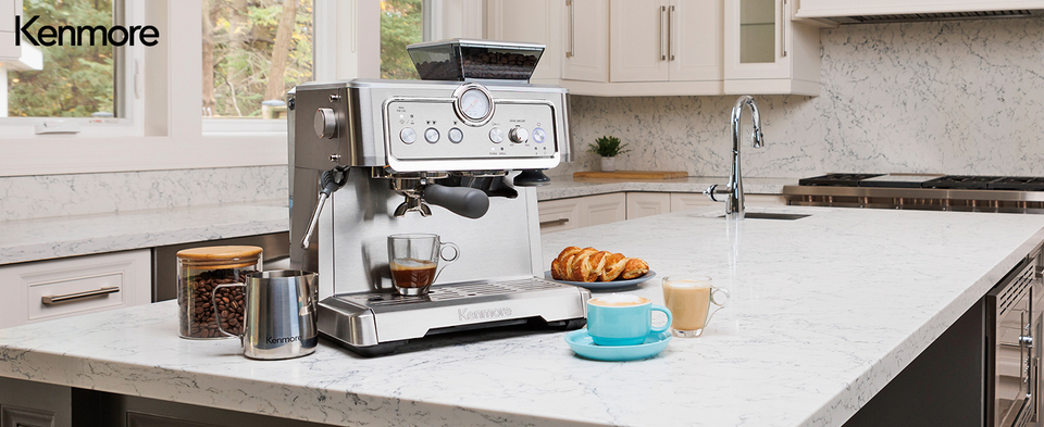 Kenmore semi-automatic espresso maker with espresso being extracted into two clear cups on a white marble countertop with the milk pitcher, a pastry, and a cappuccino in a light blue mug arranged around it.