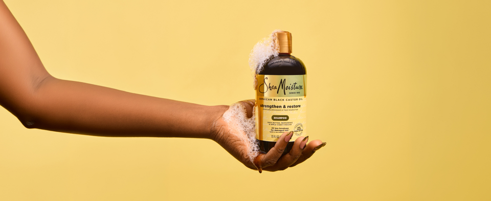 Hand holding SheaMoisture Strengthen &amp; Restore shampoo bottle with lather on gold.