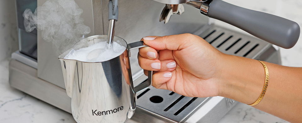 Closeup image of a person&#39;s hand holding the stainless steel pitcher filled with foamy milk with the steam wand immersed in it